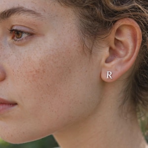 May include: A small, silver-coloured stud earring shaped like the capital letter R. The earring is positioned on the earlobe, featuring a polished metallic finish and a simple, clean serif font design that sits flush against the skin.