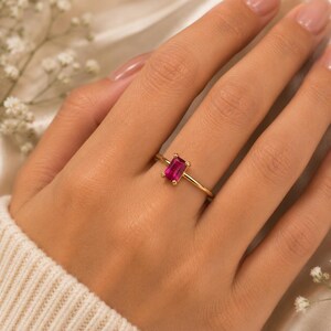 May include: A gold ring featuring a single rectangular baguette-cut vibrant pink gemstone set in a four-prong setting on a thin, polished gold band. The ring is positioned on a finger, showcasing the clear, faceted pink stone and the gold metal.