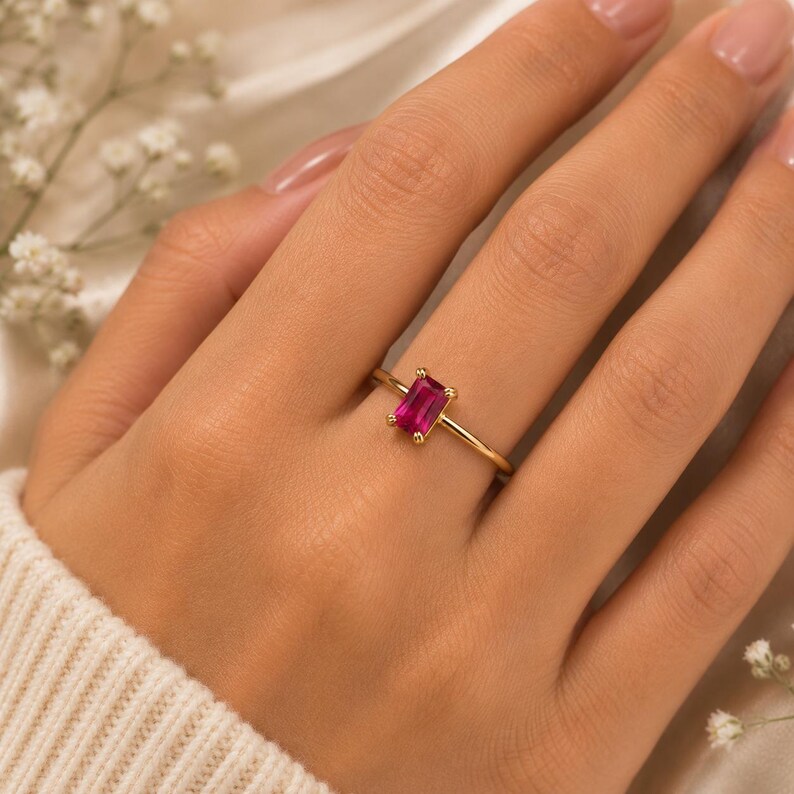 May include: A gold ring featuring a single rectangular baguette-cut vibrant pink gemstone set in a four-prong setting on a thin, polished gold band. The ring is positioned on a finger, showcasing the clear, faceted pink stone and the gold metal.
