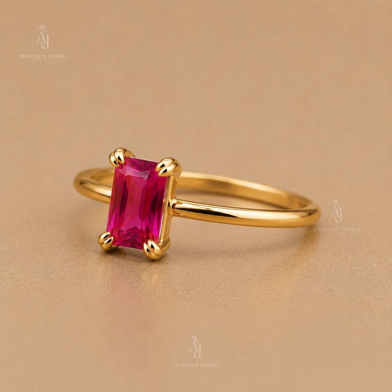 May include: A gold ring featuring a rectangular emerald-cut vibrant pink gemstone set in a four-prong setting. The band is thin, polished, and smooth, showcasing a minimalist design that highlights the bright color of the central stone.
