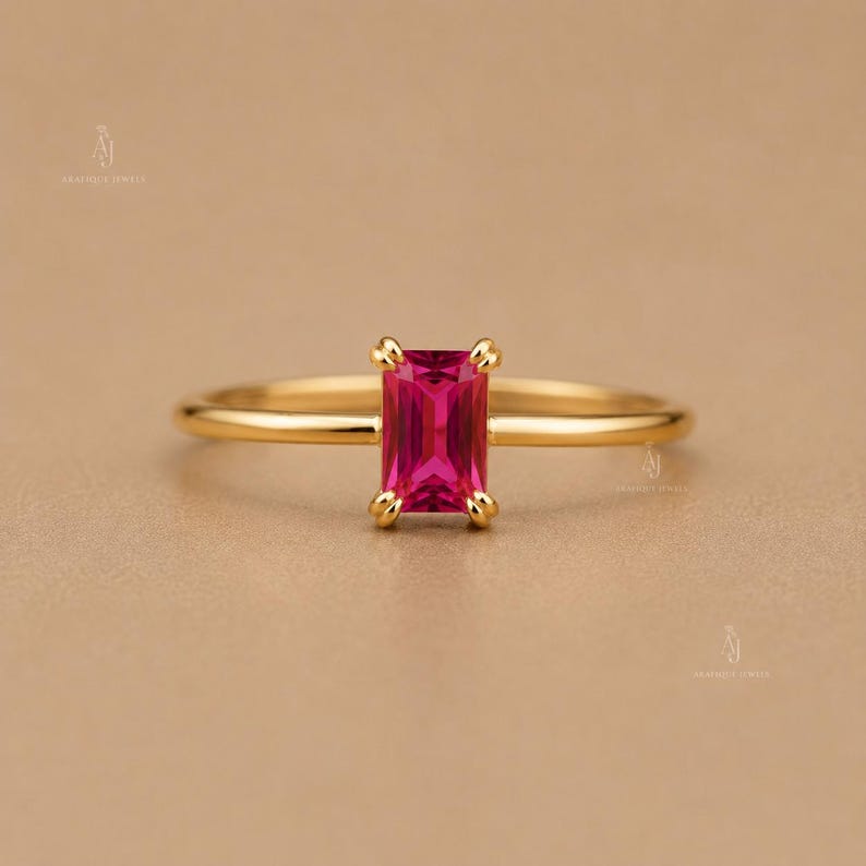 May include: A gold ring featuring a rectangular emerald-cut vibrant pink gemstone set in a four-prong setting. The band is thin, smooth, and polished, showcasing a minimalist design that highlights the central stone's deep color and clarity.