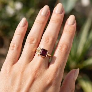 Könnte beinhalten: Ein goldener Ring mit einem quadratisch geschliffenen, tiefroten Edelstein in einer Zargenfassung. Die Ringschiene ist an einer Seite mit einer Gruppe aus drei kleinen, detaillierten goldenen Blättern verziert. Der Ring wird an einem Finger getragen.