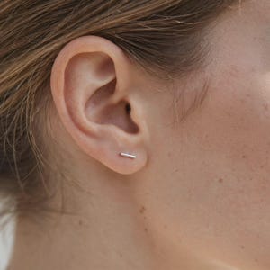 Könnte beinhalten: Ein kleiner, silberfarbener Stab-Ohrstecker, der im Ohrläppchen getragen wird. Der Ohrstecker hat ein schlichtes, horizontales, zylindrisches Metalldesign mit einer polierten, reflektierenden Oberfläche, die flach auf der Haut aufliegt.