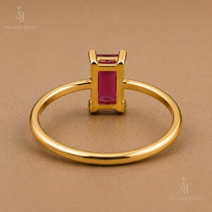 May include: A gold ring featuring a rectangular, emerald-cut deep pink gemstone set in a four-prong setting. The band is thin, polished, and circular, showcasing a minimalist design that highlights the vibrant color of the central stone.