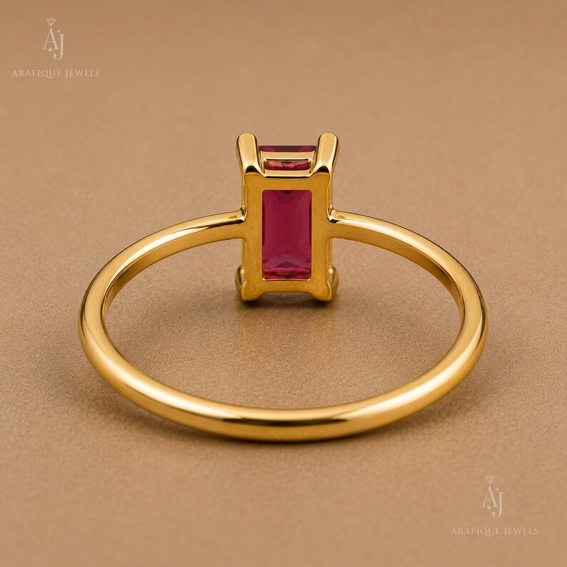 May include: A gold ring featuring a rectangular, emerald-cut deep pink gemstone set in a four-prong setting. The band is thin, polished, and circular, showcasing a minimalist design that highlights the vibrant color of the central stone.