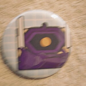 Peut inclure: Un badge rond à épingle présentant une illustration graphique d'un objet rectangulaire violet ressemblant à un livre avec un cercle jaune au centre. Le fond du motif se compose d'un quadrillage bleu clair et l'image est légèrement floue.