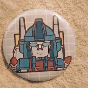 May include: A circular pinback button featuring a graphic illustration of a blue and red robot character with a helmet and visor. The character is set against a light blue background with a white grid pattern, rendered in a clean, bold line art style.