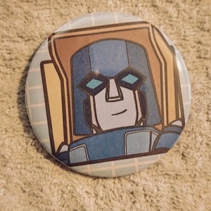 May include: A circular pinback button featuring a graphic illustration of a blue and tan robot character with geometric features, teal-colored eyes, and a light blue grid pattern background. The design is centered on the round button surface.