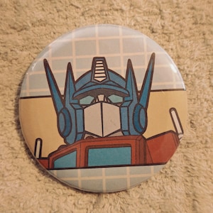 May include: A circular pinback button featuring a graphic illustration of a blue and red robot head with a pointed helmet design. The background consists of a light blue grid pattern over a tan horizontal band, creating a retro aesthetic.