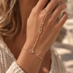 Puede incluir: Una pulsera de mano de oro con eslabones delicados y dos dijes ovalados incrustados con pequeñas piedras blancas. La cadena se extiende desde la muñeca hasta un anillo, con piedras pequeñas adicionales engastadas a lo largo de los eslabones.
