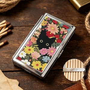 以下が含まれることがあります： 赤、黄、ピンク、白の花々が描かれたカラフルな花柄から黒猫が顔を覗かせているデザインの長方形の銀色金属製ケース。円形のインセットには、Harrietという名前が刻印された金属製のバーが表示されています。