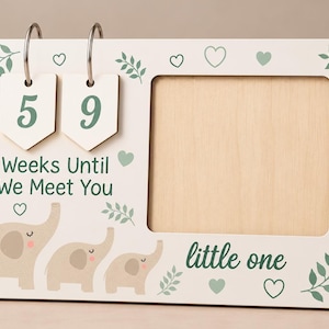 Pode incluir: Uma placa retangular de madeira para contagem regressiva com duas etiquetas penduradas exibindo 5 e 9, o texto "Weeks Until We Meet You" e "little one", decorada com folhas verdes, pequenos corações e três elefantes bege.