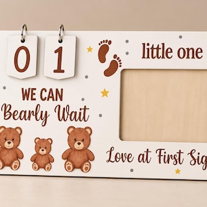 Könnte beinhalten: Ein weißer, rechteckiger Countdown-Rahmen aus Holz mit zwei umklappbaren Zahlenkarten, die 01 zeigen, sowie Illustrationen von braunen Bären und Babyfußabdrücken. Der Text lautet: We Can Bearly Wait, little one, und Love at First Sight, verziert mit gelben Sternen.