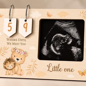 May include: A light-coloured wooden pregnancy countdown frame featuring two hanging number tags displaying 5 and 9, the text "WEEKS UNTIL WE MEET YOU" and "Little one", a square photo opening, and illustrations of a bear, a lion, butterflies, and clouds.