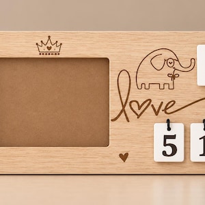 May include: A rectangular light wood photo frame featuring a laser-engraved crown, hearts, an elephant, and the word love. The frame includes three white hanging tiles displaying the text TUE, 5, and 1, suspended by black cords from the wood.