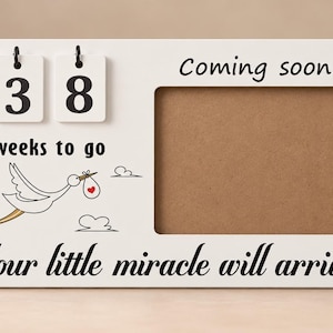 Könnte beinhalten: Ein weißer, rechteckiger Bilderrahmen aus Holz mit einem Countdown-Display aus zwei Klappkarten, die die Zahl 38 zeigen. Der Rahmen trägt die Aufschriften Coming soon, weeks to go und Your little miracle will arrive sowie eine Storch-Grafik.