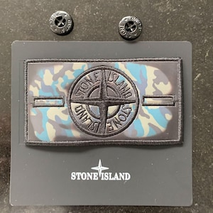 Könnte beinhalten: Ein rechteckiger Stone Island Aufnäher mit blau-grau-schwarzem Tarnmuster und einem zentralen, gestickten Kompass-Logo. Der Aufnäher hat zwei Knopflöcher und wird von zwei schwarzen, runden Knöpfen mit Stone Island Schriftzug begleitet.