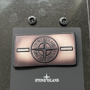 May include: A rectangular Stone Island patch featuring a central compass logo with black embroidery on a gradient brown background. The patch includes two horizontal buttonholes and is accompanied by two black buttons and a branded display card.