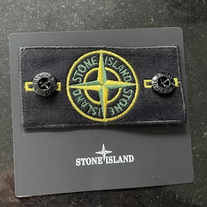 Pode incluir: Um emblema retangular de tecido preto com o logótipo da bússola Stone Island bordado a amarelo e verde. O emblema inclui dois botões pretos com o texto e o logótipo Stone Island, montados num cartão de apresentação preto com a marca.
