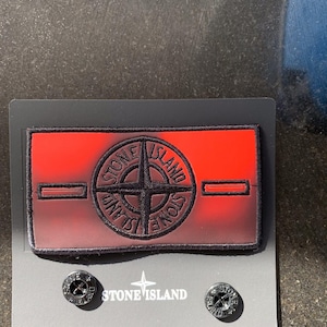 Puede incluir: Un parche rectangular con degradado rojo y negro que presenta el logotipo de la brújula de Stone Island bordado y dos ranuras horizontales. Debajo del parche, dos botones metálicos circulares están fijados a la tarjeta, grabados con el logo.