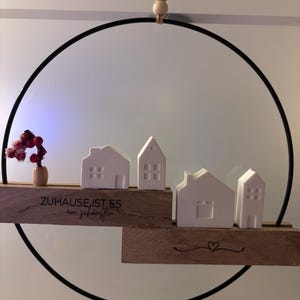 Könnte beinhalten: Ein schwarzer Metallring, der an einer Jutekordel mit drei Holzperlen hängt. Zwei Holzblöcke tragen vier weiße Keramikhausfiguren und einen kleinen roten Blumenzweig. Das Holz ist mit dem Text "ZUHAUSE IST ES am schönsten" und einem Herz graviert.