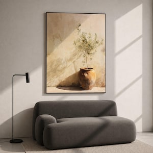 Op de afbeelding: Een ingelijste kunstdruk met een kleine olijfboom in een rustieke, verweerde terracotta pot. Het kunstwerk toont een warm, neutraal kleurenpalet met zacht zonlicht dat lange schaduwen werpt tegen een gestructureerde, beige gepleisterde muur.