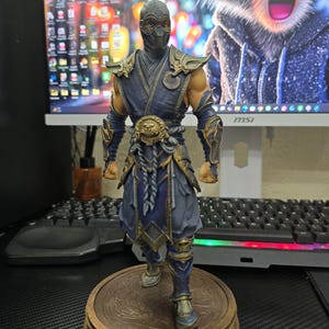 May include: A standing figurine of a ninja character wearing a dark blue and gold armored outfit with a matching mask. The figure features intricate shoulder guards, a detailed belt buckle, and stands on a circular, textured bronze-colored base.