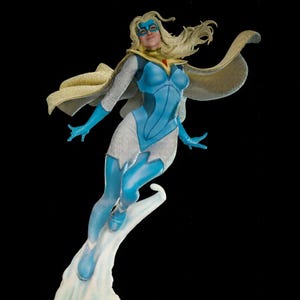 Op de afbeelding: Een superheldenfiguur met lang blond haar, een blauw masker en een blauw-wit pak. Het figuur draagt een gouden cape met een rood embleem bij de nek en is afgebeeld in een dynamische vlieghouding op een witte wolk als basis.