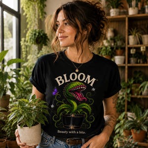 May include: A black t-shirt featuring a graphic of a green Venus flytrap in a black pot with the text "BLOOM" above and "Beauty with a bite." below. The design includes a small purple butterfly and white insects surrounding the plant.
