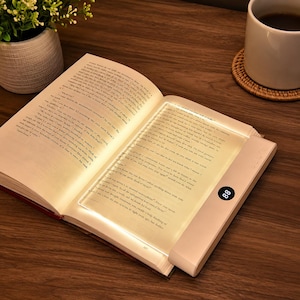 May include: A rectangular, flat, white LED book light panel resting on an open book. The device features a circular digital display showing the number 88 and a side-mounted control panel. The light illuminates the pages of the open book.