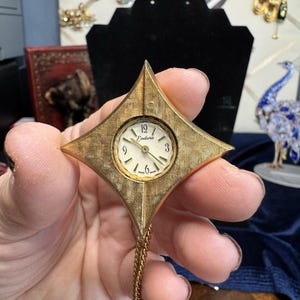 Peut inclure: Une montre pendentif dorée avec un boîtier en forme de losange à la surface texturée. Le cadran rond blanc affiche le nom de la marque Endura, des chiffres noirs aux positions 3, 6, 9 et 12, et des aiguilles dorées. Une chaîne dorée est fixée en bas.