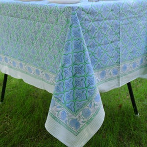 Puede incluir: Un mantel azul claro y verde con un patrón floral repetitivo dentro de una cuadrícula de líneas festoneadas. La tela tiene un borde ancho con un diseño de rayas y un motivo floral, que cuelga sobre el borde de una mesa.