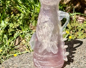 May include: A translucent glass pitcher with a tapered neck and a wide base, featuring a pale lavender and white mottled finish. The vessel has a curved handle on one side and a series of small, decorative glass protrusions along the outer edge.
