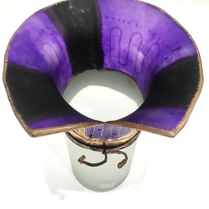 May include: A decorative vase with a wide, flared rim. The interior is divided into purple and black sections. The rim has a brown edge. The vase sits atop a frosted glass container with a decorative rope detail.