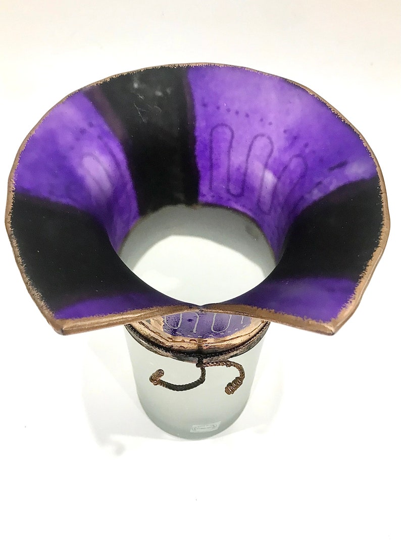 May include: A decorative vase with a wide, flared rim. The interior is divided into purple and black sections. The rim has a brown edge. The vase sits atop a frosted glass container with a decorative rope detail.