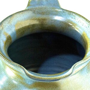 May include: A ceramic pitcher featuring a wide, rounded opening with a small pouring spout. The exterior surface displays a mottled glaze with blended tones of sage green, earthy brown, and soft blue, creating a textured, organic appearance.