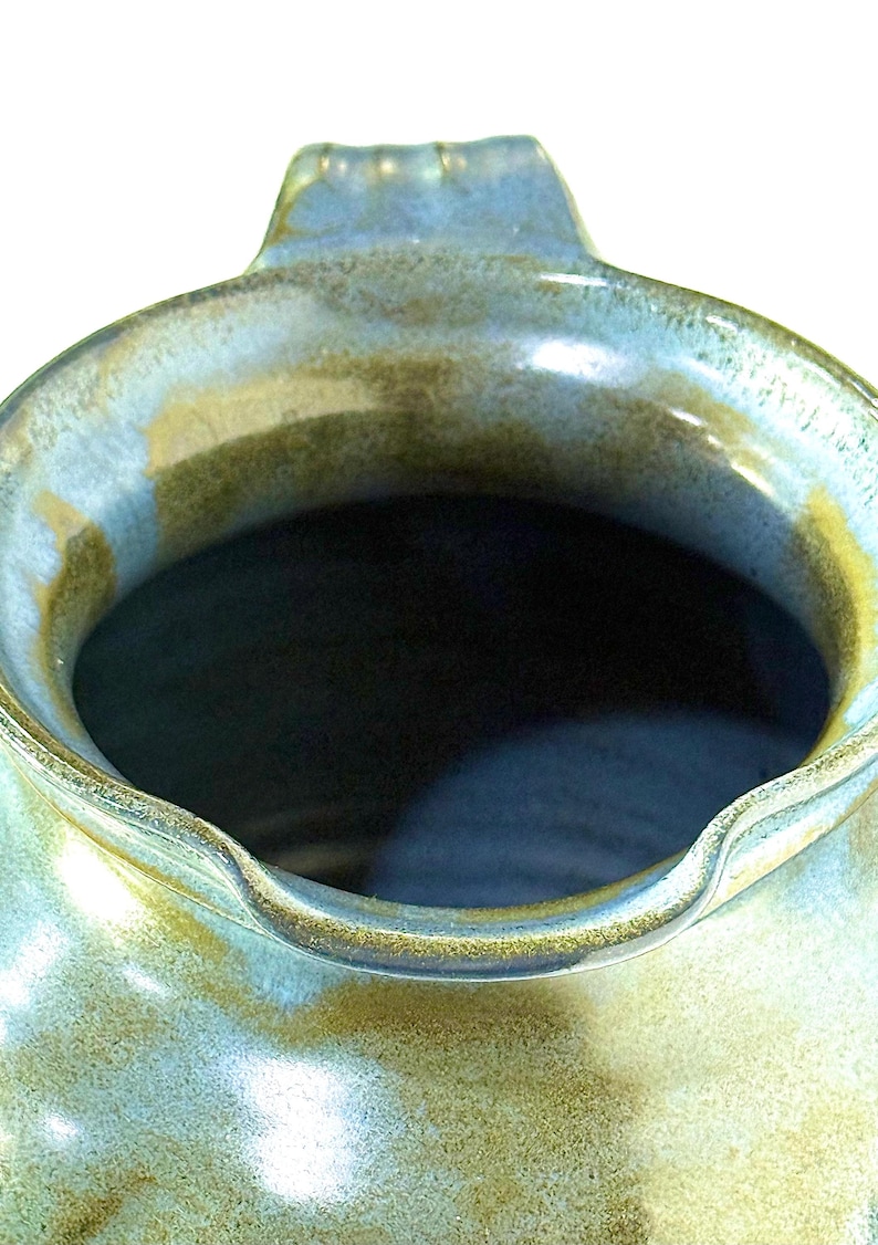 May include: A ceramic pitcher featuring a wide, rounded opening with a small pouring spout. The exterior surface displays a mottled glaze with blended tones of sage green, earthy brown, and soft blue, creating a textured, organic appearance.
