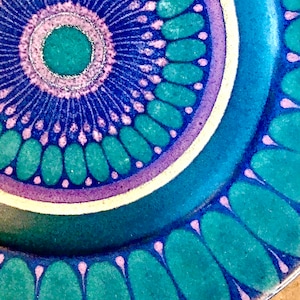 May include: Close-up of a ceramic plate with a detailed, colorful design. The plate features a central teal circle surrounded by concentric rings of blue, purple, and teal, with a white border. The outer edge has a repeating pattern of teal shapes.