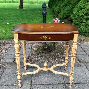 May include: A rectangular side table with a dark brown wooden top featuring a central floral painting. The table has four cream-colored, distressed, fluted legs connected by a cross-shaped stretcher with a small finial in the center.