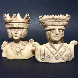May include: Two off-white ceramic busts of crowned figures. The figures have detailed facial features and wear ornate crowns. The busts have decorative shoulder pieces. The background is a dark blue-gray.
