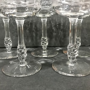 May include: A close-up of five clear champagne glasses. The glasses have a round base, a twisted stem, and a bowl with a decorative etched pattern. The glasses are arranged in a row, with the stems and bases visible.