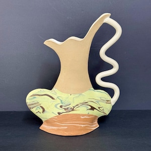 May include: A ceramic pitcher featuring a tall, ribbed beige neck and a wide, flared base. The base displays a marbled pattern of light green, brown, and cream. A distinct, white, serpentine-shaped handle extends from the side to the top rim.