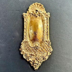 May include: Ornate gold-colored decorative plaque with a rectangular center and intricate floral and geometric patterns. The plaque features a sunburst design at the top and is likely made of metal. The overall design is vintage and detailed.