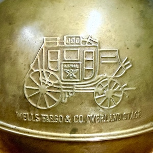 May include: A polished brass object featuring an embossed graphic of a stagecoach with the text WELLS FARGO & CO. OVERLAND STAGE. The metal surface has a warm, metallic gold tone with visible engraved details of the carriage wheels and frame.