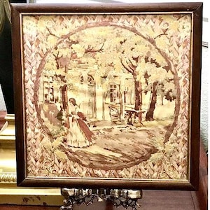 May include: A framed artwork depicting a scene with a woman in a long dress, trees, and a building. The artwork is in a brown wooden frame and is displayed on a decorative metal stand. The color palette includes shades of brown, beige, and white.