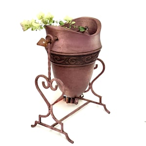 May include: A mauve-colored metal vessel with a textured finish, featuring a decorative band with embossed scrollwork. The vessel is suspended within a matching metal stand that has heart-shaped side supports and a small sprig of greenery.