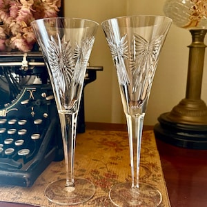 May include: Two clear crystal champagne flutes featuring a tall, slender stem and a wide, circular base. The upper portion of each glass is decorated with intricate, deep-cut starburst and fan patterns that catch the light, creating a reflective effect.