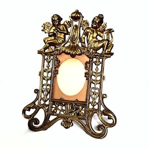 May include: Ornate brass-toned picture frame featuring two cherubic figures at the top. The frame has an oval opening and decorative floral and scrollwork details. The frame is approximately 8 inches tall.