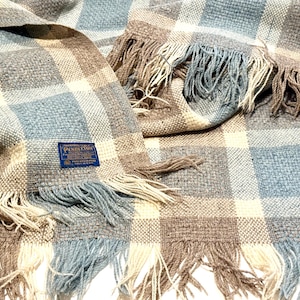 May include: A woven blanket with a plaid pattern in shades of gray, blue, beige, and brown. The blanket has fringed edges and a blue label with white text. The blanket is made of a soft, textured material.