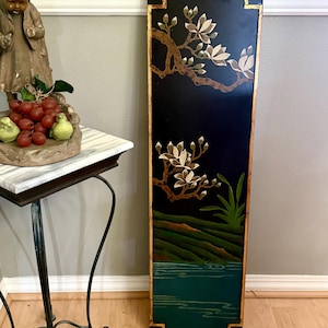 May include: A tall, rectangular black panel with a gold border and floral design. The artwork features a tree branch with white and brown flowers, green foliage, and a landscape scene with water.